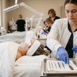A student esthetician wearing blue gloves prepares a professional skincare tray with labeled bottles and metal tools in a bright classroom clinic.