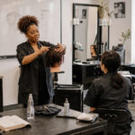 Beauty instructor demonstrating haircutting technique on a mannequin head while adult students observe and take notes in a professional cosmetology training classroom.