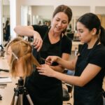 An experienced beauty educator demonstrating professional hair sectioning and cutting on a mannequin head while two adult students observe and take notes in a realistic classroom setting.