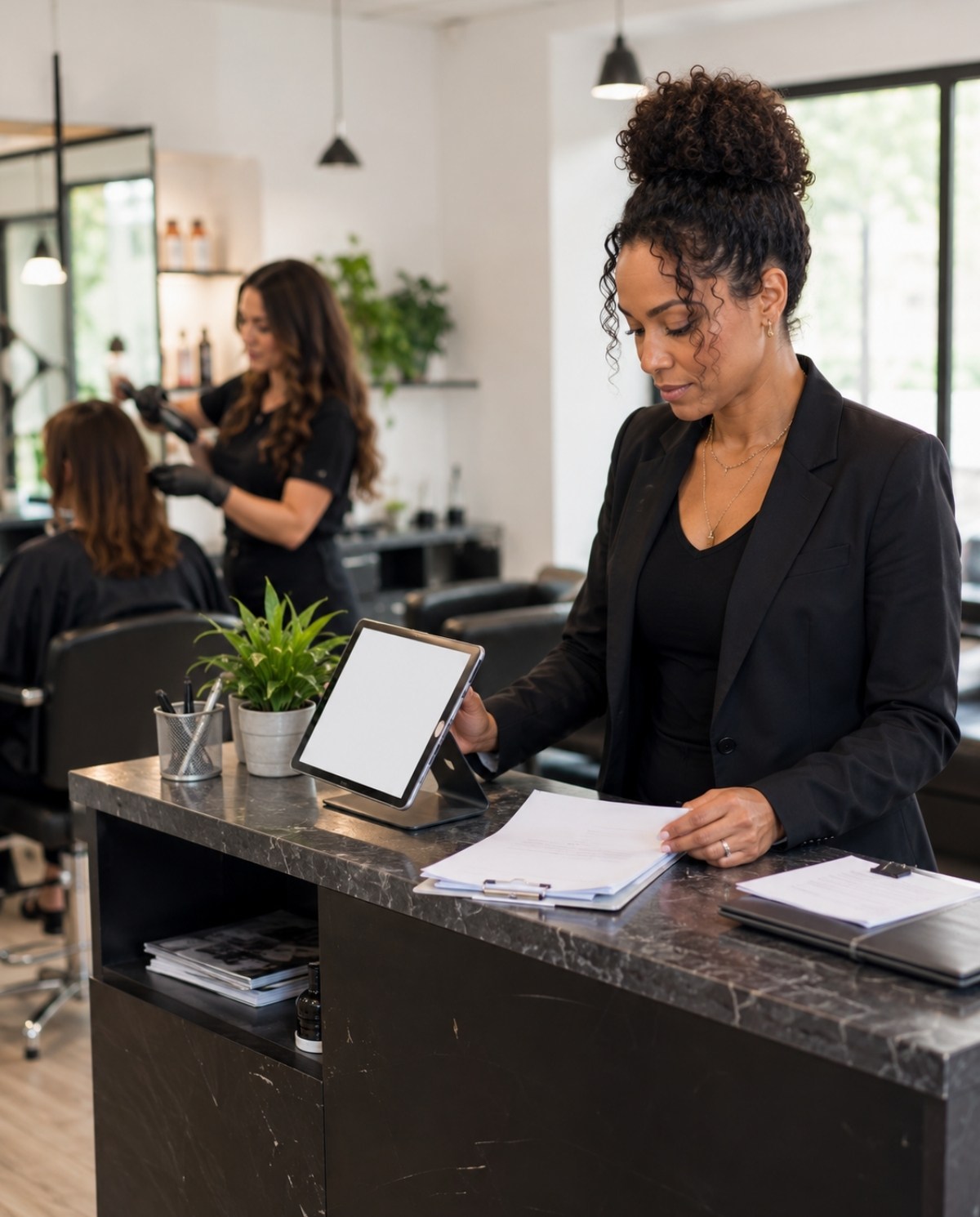 Salon business owner checking a blank appointment tablet at the reception counter while a licensed professional works at a styling chair in the background.