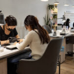 Professional nail salon with multiple manicure stations featuring supportive seating, task lamps, organized supplies, practical worktables, and source-capture ventilation near each service area.