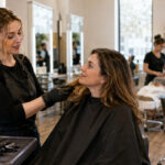 Cosmetology student examining a client’s hair texture during a consultation while manicure and facial training take place at other stations in the background.