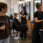 Prospective beauty student carrying a notebook while observing student stylists consulting with clients during hands-on cosmetology training.