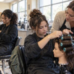 High school students practicing hair roller placement on mannequin heads while an instructor explains proper section sizing.