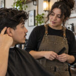 A client with thick, wavy dark hair points just above his ear while a female barber listens with a comb in her hands.